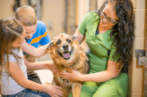 Woman in green scrubs kneeling next to two children. All are petting a brown dog in a humane society.